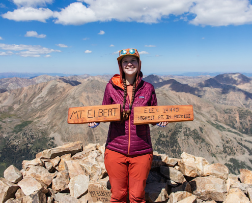 Emma standing on the summit of Mount Elbert holding wooden sign at 14,440 feet, highest 14er in Colorado Rockies