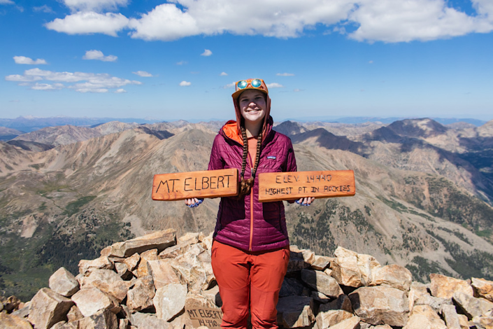 Emma standing on the summit of Mount Elbert holding wooden sign at 14,440 feet, highest 14er in Colorado Rockies