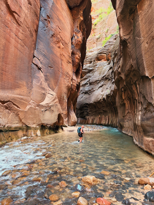 Distant hiker walking through the Virgin River beneath towering canyon walls during The Narrows hike in Zion National Park.