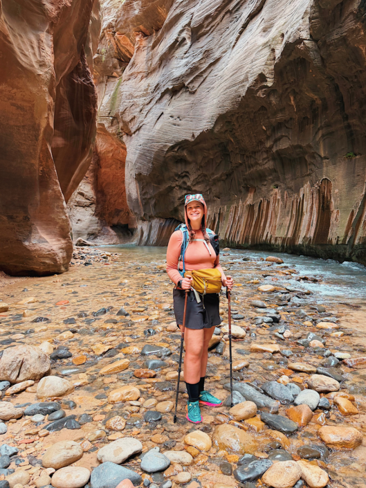 Close-up of a hiker standing in the river with trekking poles during The Narrows hike, backed by a curved sandstone canyon wall in Zion National Park.