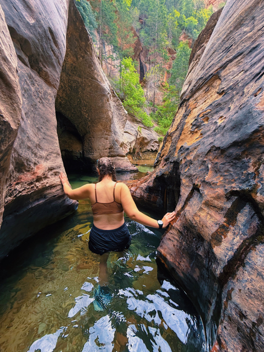 Hiker walking through narrow sandstone potholes and holding canyon walls for balance on the Zion Subway hike