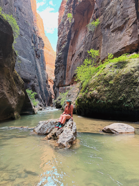 Sitting on a rock in the Virgin River while hiking the Narrows in Zion National Park with towering canyon walls behind me during a Zion vs Bryce National Park trip.