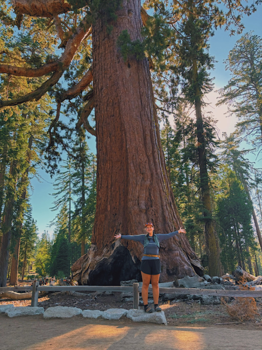 Hiker standing with arms out in front of the massive Grizzly Giant in Mariposa Grove, Yosemite