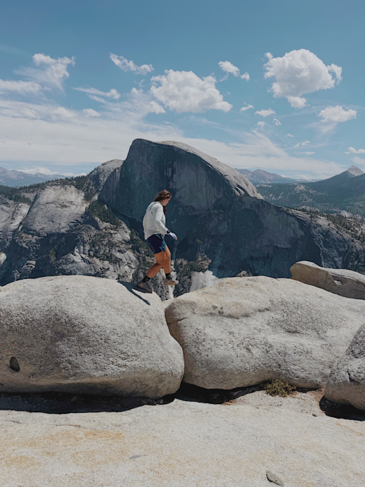 Hiker walking across granite rocks with Half Dome in the background on the North Dome Yosemite hike