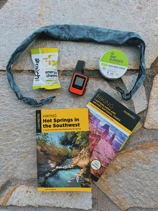 Hiking guidebooks, Garmin inReach, sunscreen, energy chews, and cooling neck tie arranged on a rock