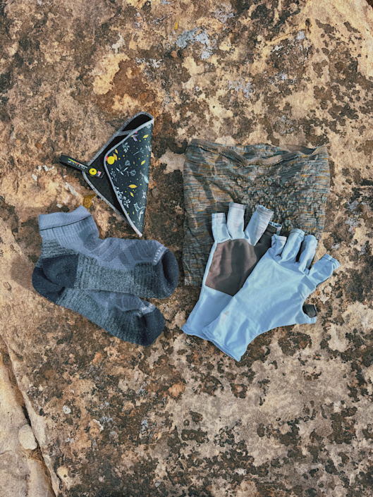 Merino wool hiking socks, Kula Cloth, neck gaiter, and sun gloves arranged on a rock