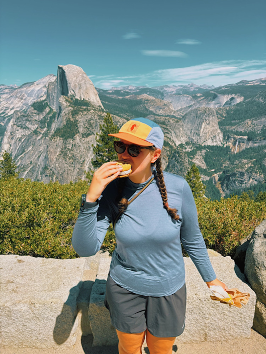 Woman enjoying an ice cream sandwich at Glacier Point with Half Dome and the Sierra Nevada mountains behind her.