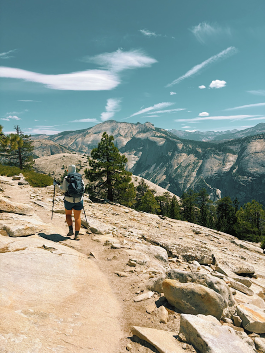 Hiker walking across granite terrain on the North Dome Trail with expansive views of Yosemite's high country.