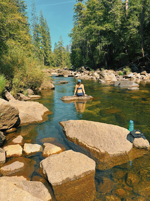 Woman sitting on a large boulder in the shallow Merced River, surrounded by clear water, rocks, and pine trees.
