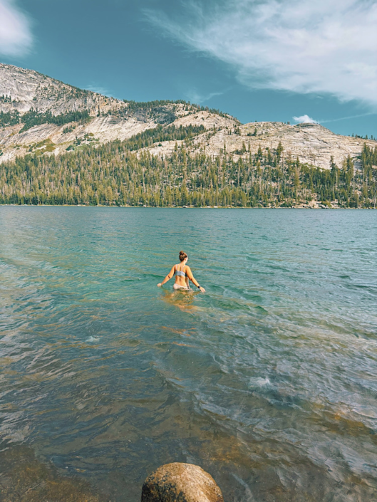 Woman wading into the blue water of Tenaya Lake with granite mountains and pine forests in the background.
