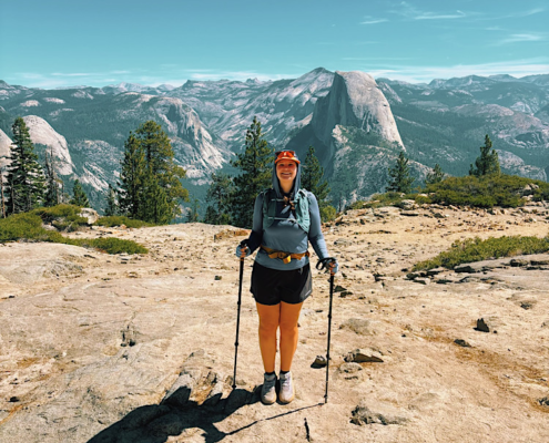 Hiker standing on Sentinel Dome with trekking poles and Half Dome rising above the mountains in the background.