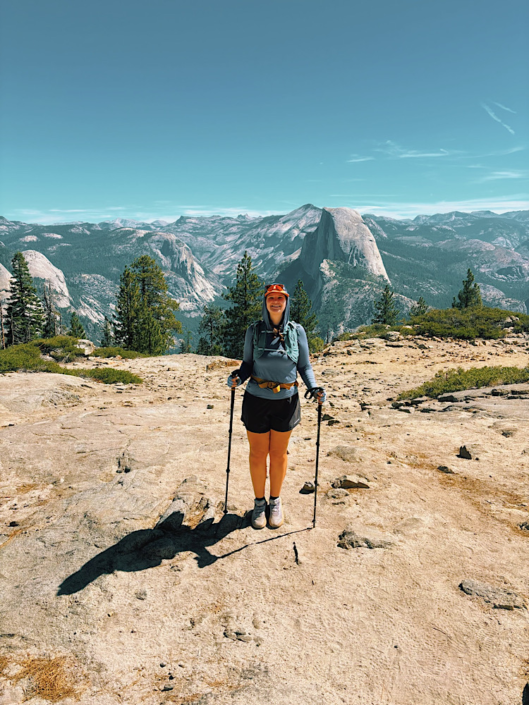 Hiker standing on Sentinel Dome with trekking poles and Half Dome rising above the mountains in the background.
